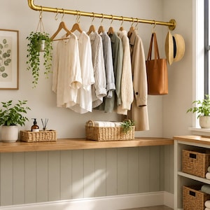 Puede incluir: Un perchero de metal dorado montado en el techo y la pared, con múltiples ganchos en S que sostienen perchas de madera con varias camisas, un abrigo largo, un bolso de cuero marrón, un sombrero de paja y una planta colgante.