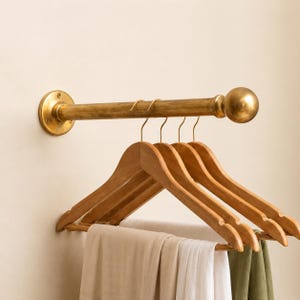 Könnte beinhalten: Eine wandmontierte Kleiderstange aus Messing mit einer kugelförmigen Endkappe und einer gebürsteten Metalloberfläche. Drei Holzkleiderbügel hängen an der horizontalen Stange, an denen gefaltete Stoffe in Beige, Weiß und Olivgrün hängen.
