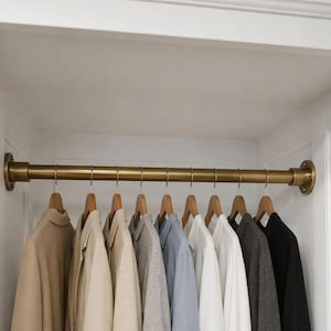 Puede incluir: Una barra de armario de metal en tono latón montada entre dos paredes blancas, con bridas de pared circulares en cada extremo. Nueve perchas de madera cuelgan de la barra, cada una con una prenda en tonos neutros que incluyen beige, gris, azul, blanco y negro.