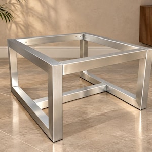 Puede incluir: Una base de mesa de centro cuadrada fabricada en metal plateado cepillado. El marco cuenta con cuatro patas verticales conectadas por barras horizontales en la parte superior e inferior, creando una estructura geométrica abierta de diseño industrial.