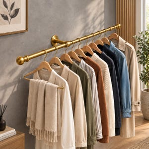 Pode incluir: Um cabideiro de parede em metal dourado com uma barra horizontal e extremidades esféricas. Vários cabides de madeira estão suspensos na barra, segurando diversas peças de vestuário, incluindo um cachecol bege, camisas, suéteres e um sobretudo.