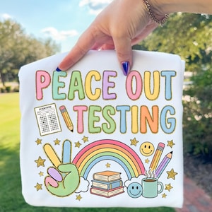 Op de afbeelding: Wit t-shirt met de tekst "PEACE OUT TESTING" in kleurrijke letters. Het ontwerp bevat een vredesteken, een regenboog, boeken, potloden, een koffiekopje en sterren. Een Scantron-formulier is ook inbegrepen.