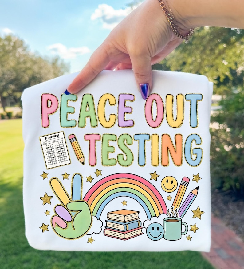 Op de afbeelding: Wit t-shirt met de tekst "PEACE OUT TESTING" in kleurrijke letters. Het ontwerp bevat een vredesteken, een regenboog, boeken, potloden, een koffiekopje en sterren. Een Scantron-formulier is ook inbegrepen.