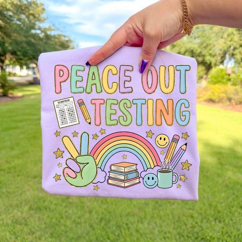 Op de afbeelding: Een lichtpaars t-shirt met de tekst "PEACE OUT TESTING" in kleurrijke letters. Het ontwerp bevat een vredesteken, een regenboog, boeken, potloden, sterren, een smiley en een koffiekopje.