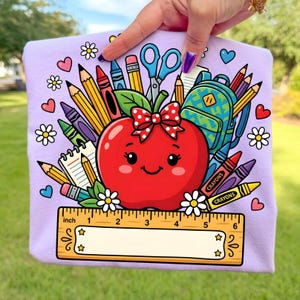 Puede incluir: Camiseta de color lila claro con una manzana de dibujos animados con un lazo rojo, rodeada de útiles escolares como lápices, crayones y una mochila. Una regla con medidas en pulgadas en la parte inferior. Tema de regreso a clases.