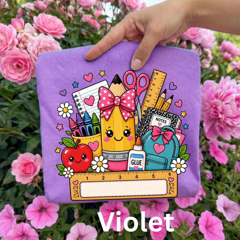 May include: A light purple t-shirt featuring a colorful graphic of a smiling yellow pencil with a pink polka dot bow, a red apple, a blue backpack, scissors, glue, and a ruler with the text "NOTES" on a notebook and "GLUE" on a bottle.