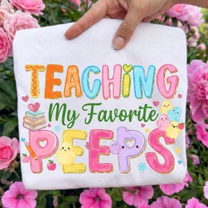 May include: White t-shirt with the text "TEACHING My Favorite PEEPS" in colorful letters. The design includes hearts, books, a pencil, and cartoon characters. The shirt is held up by a hand, surrounded by pink flowers.