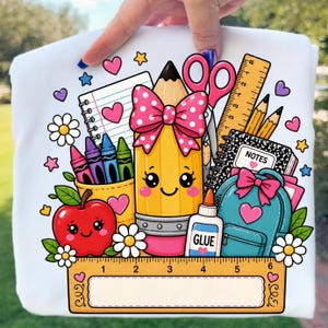 May include: A white t-shirt featuring a colorful graphic of school supplies including a smiling yellow pencil with a pink polka dot bow, a red apple, a blue backpack, scissors, glue, crayons, a notebook, and a ruler with 6-inch markings.