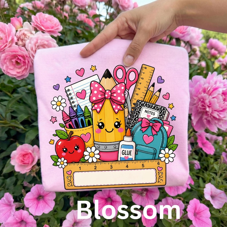 May include: A light pink t-shirt featuring a colorful graphic of school supplies including a smiling pencil with a pink polka dot bow, a red apple, scissors, a glue bottle, a backpack, a notebook, and a ruler with a blank white label area.