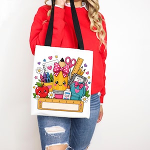 May include: A white tote bag with black straps featuring a colorful graphic of school supplies including a smiling pencil with a pink polka dot bow, a red apple, a blue backpack, a ruler, glue, and notebooks, all decorated with small hearts and flowers.
