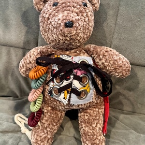 May include: A brown plush teddy bear with a black nose and eyes. The bear has a colorful fabric panel on its chest with metal eyelets and black lacing. A string of colorful hair ties and a red strap are attached to the bear.