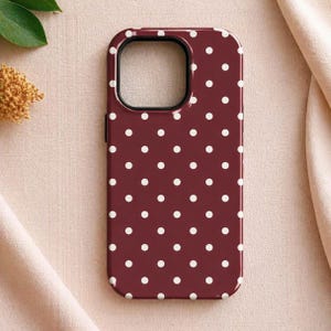 May include: A burgundy phone case with a repeating pattern of white polka dots. The case has a black border around the camera cutout and a glossy finish. The case is designed to protect a smartphone.
