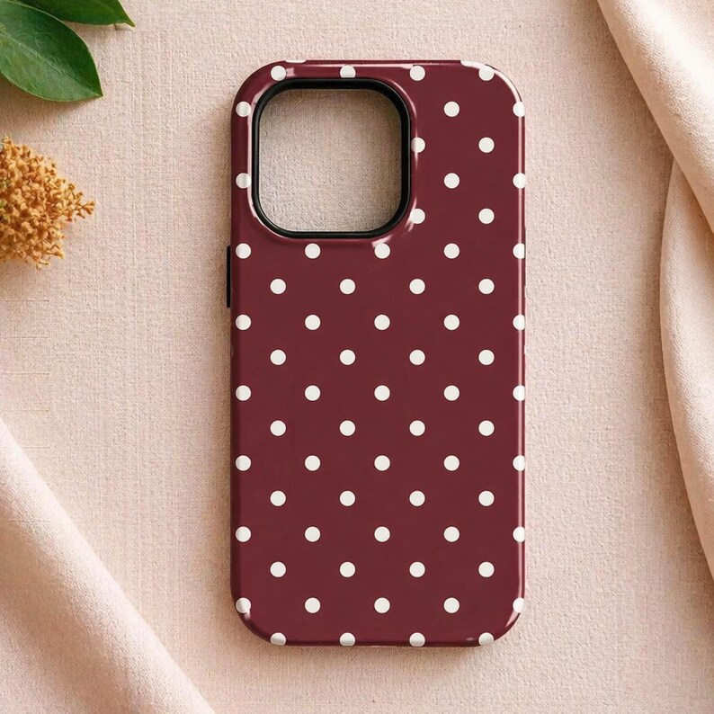 May include: A burgundy phone case with a repeating pattern of white polka dots. The case has a black border around the camera cutout and a glossy finish. The case is designed to protect a smartphone.