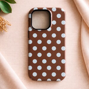May include: A brown phone case with a pattern of light blue polka dots. The case has a black border around the camera cutout. The phone case is a protective accessory for a smartphone.