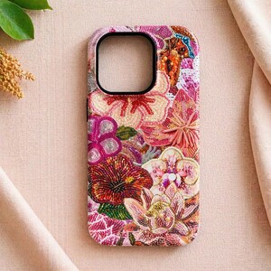 May include: A decorative phone case with an elaborate floral pattern. The case displays a vibrant mix of pink, red, and orange beaded flowers. The case has a black border around the camera cutout.