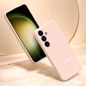 May include: A light pink phone case and a smartphone with a light green screen. The phone case has three camera lenses and the word "SAMSUNG" printed on it. The phone case is designed to protect the phone.