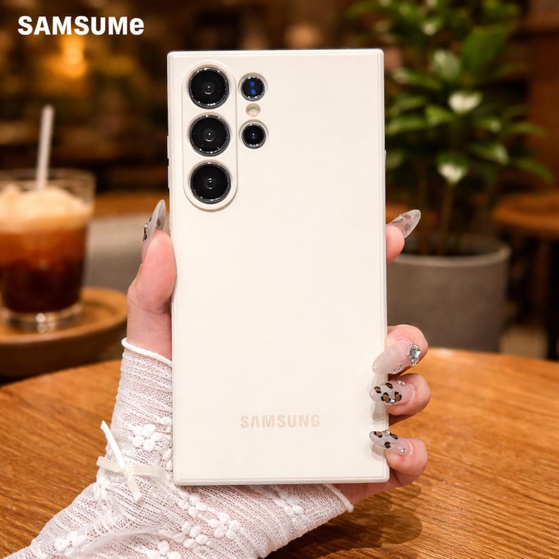 May include: A cream-colored phone case with multiple camera lenses and the Samsung logo. The case has the brand name "SAMSUNG" printed on the lower part of the case. The brand name "SAMSUME" is printed on the top left corner of the case.