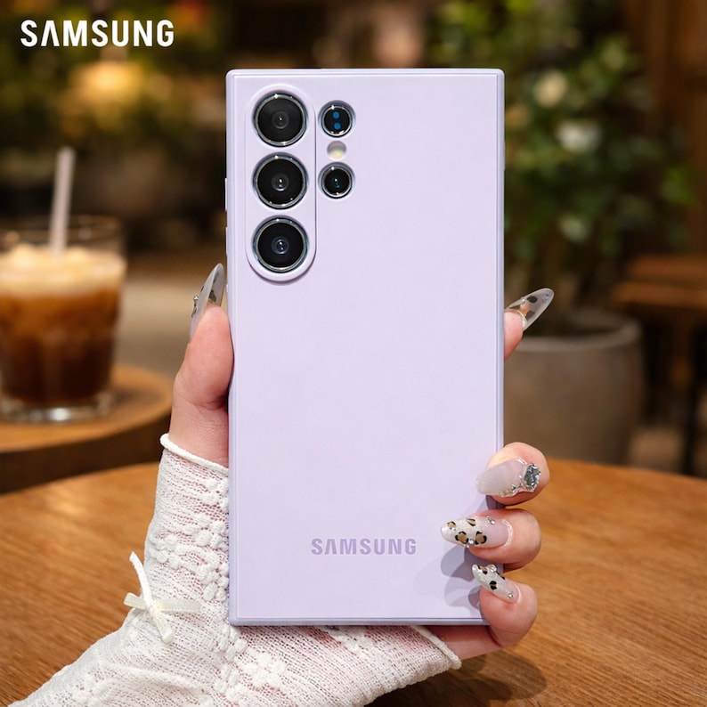 May include: A light purple Samsung phone case with multiple camera lenses. The Samsung logo is visible at the bottom. The phone case is held by a person with long nails. The background is blurred.