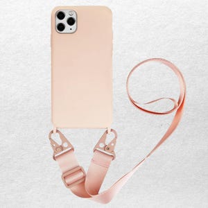 May include: A blush pink phone case with a matching strap. The case has a camera cutout and a strap that attaches with metal clasps. The strap is adjustable and can be worn around the neck or across the body. The phone case is made of a smooth, matte material.