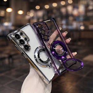 May include: Two phone cases, one silver and one purple, with a clear back. Both cases have a ring holder with the text "SATRY TECH". The silver case has a silver frame, and the purple case has a purple frame.