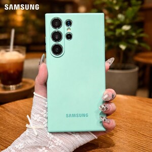 May include: A mint green Samsung phone case with multiple camera lenses. The Samsung logo is visible at the top and bottom of the case. The phone case is held by a person with long nails.