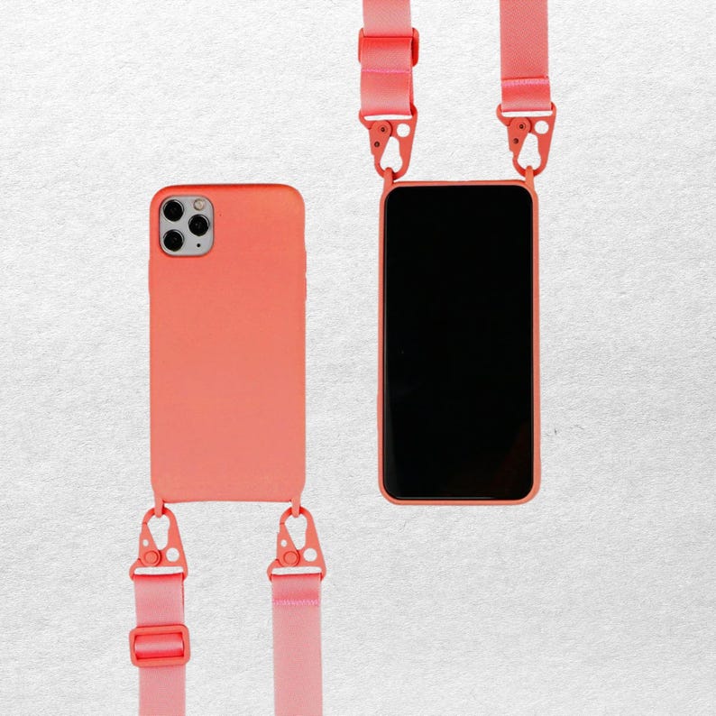 May include: A coral-colored smartphone case with a matching adjustable strap attached via two metal clips. The case features a triple-lens camera cutout on the back and a black screen on the front, providing a hands-free carrying solution for mobile devices.