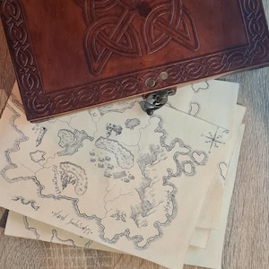 May include: A brown leather journal with Celtic knotwork on the cover sits above several parchment maps. The maps are hand-drawn with detailed illustrations of islands, coastlines, and settlements. The journal is a rich brown colour, and the maps are cream-coloured.