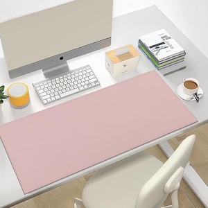 Könnte beinhalten: Eine große, rechteckige, einfarbige hellrosa Schreibtischunterlage mit genähten Kanten. Die Unterlage liegt auf einem weißen Schreibtisch vor einem Computermonitor, einer weißen Tastatur, einer hölzernen Taschentuchbox, Büchern und einer Kaffeetasse.