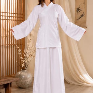 May include: A solid white two-piece outfit consisting of a long-sleeved wrap-style top with a V-neckline and wide sleeves, paired with a matching floor-length skirt. The garment features clean lines, a simple wrap closure, and a plain white fabric.