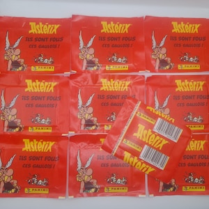 Puede incluir: Una colección de paquetes rojos de cromos de Astérix. Cada paquete presenta el logotipo de Astérix y la frase "Ils Sont Fous Ces Gaulois!" en texto amarillo. Los paquetes están dispuestos en una caja blanca.