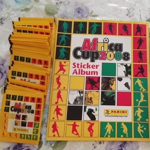 May include: A selection of unopened yellow sticker packets and an album for the Africa Cup 2008. The album cover displays the text "Africa Cup 2008 Sticker Album" with colourful illustrations of footballers and a football. The Panini logo is visible.
