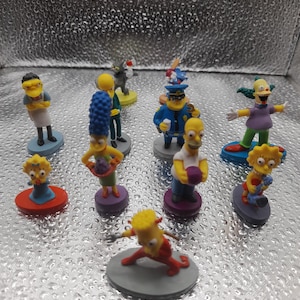 Puede incluir: Una colección de figuras coloridas con personajes de la serie de televisión animada Los Simpson. Las figuras representan personajes como Homer, Marge, Bart, Lisa y otros, cada uno de pie sobre una pequeña base redonda. Las figuras son de plástico y están pintadas con colores brillantes.