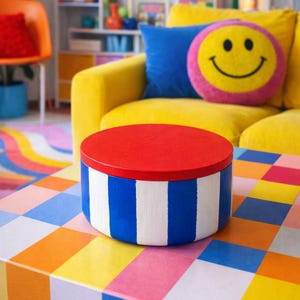 May include: A round, colorful storage box with a red lid and blue and white striped sides. The box sits on a multi-colored checkered table. The background includes a yellow sofa, a blue pillow, and a smiley face pillow.