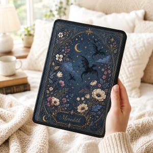 Op de afbeelding: Een rechthoekige e-readerhoes met een donkerblauwe achtergrond, voorzien van een gouden maansikkel, sterren en silhouetten van vliegende draken. Het ontwerp bevat witte en roze bloemen en de naam Meredith in gouden letters.