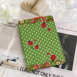 Puede incluir: Una funda rectangular verde para tableta con un patrón de lunares blancos e ilustraciones de varias mariquitas rojas. Los bordes superior e inferior están decorados con un borde de motivos florales rojos y verdes.