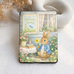 Può includere: Una custodia nera con un'illustrazione affascinante di un coniglietto che tiene in mano un fiore giallo. La scena include un tavolo con libri, fiori e la scritta "Welcome Spring". L'opera d'arte è ambientata su uno sfondo di una casa blu e una finestra con tende.