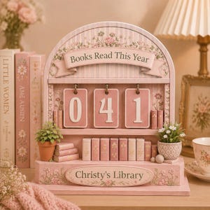 May include: A pink wooden bookshelf with the text "Books Read This Year" and "Christy's Library." The bookshelf displays the numbers "041" on pink cards. Miniature books and potted plants decorate the shelves, with floral accents.