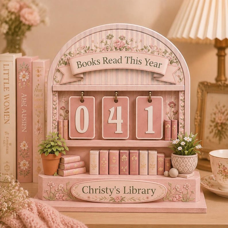May include: A pink wooden bookshelf with the text "Books Read This Year" and "Christy's Library." The bookshelf displays the numbers "041" on pink cards. Miniature books and potted plants decorate the shelves, with floral accents.