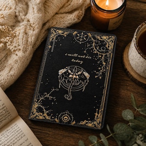 Puede incluir: Diario negro con detalles dorados y el texto "i will not die today." La portada presenta un diseño ornamentado con una ilustración central de una criatura parecida a un dragón. El diario está sobre una superficie de madera.