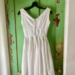 May include: A sleeveless white dress featuring a V-neckline, a fitted bodice with subtle white floral embroidery at the waist, and a pleated skirt that falls to the knee. The dress has a central vertical button closure down the front bodice.