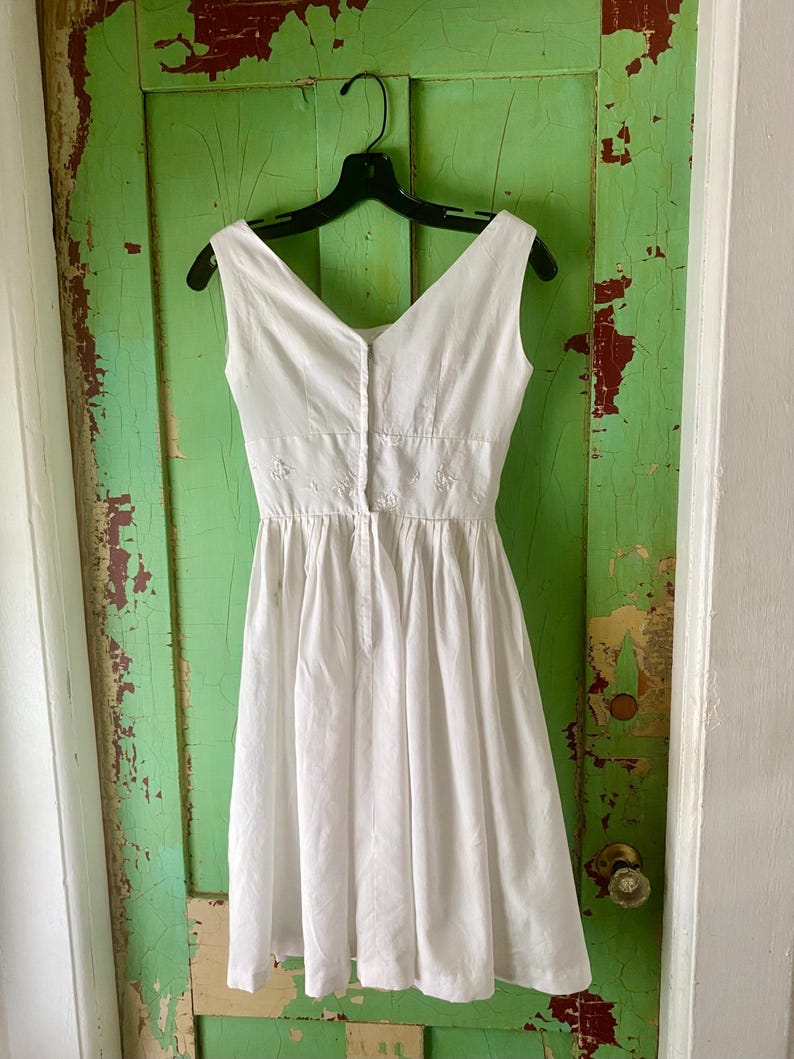 May include: A sleeveless white dress featuring a V-neckline, a fitted bodice with subtle white floral embroidery at the waist, and a pleated skirt that falls to the knee. The dress has a central vertical button closure down the front bodice.