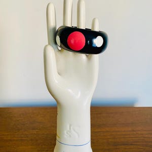 May include: A black bangle bracelet with a red and white polka dot design. The bracelet is displayed on a white ceramic hand sculpture. The sculpture is on a wooden surface.