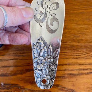 May include: Silver-toned shoe horn with an engraved floral design and a decorative initial. The handle features a detailed relief of flowers and foliage. The shoe horn is approximately 7 inches long.