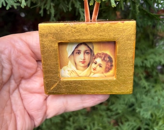 May include: A rectangular gold-leaf frame containing a small print of a woman with a white head covering holding a child. The frame has a textured, metallic finish and is suspended by a thin, light brown velvet ribbon for hanging.