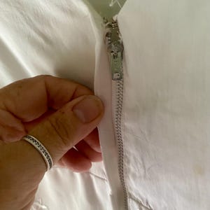 May include: A white garment featuring a vertical metal zip closure with a silver-toned pull tab. The fabric is a solid white material with subtle embroidered details visible near the bottom edge of the closure area.