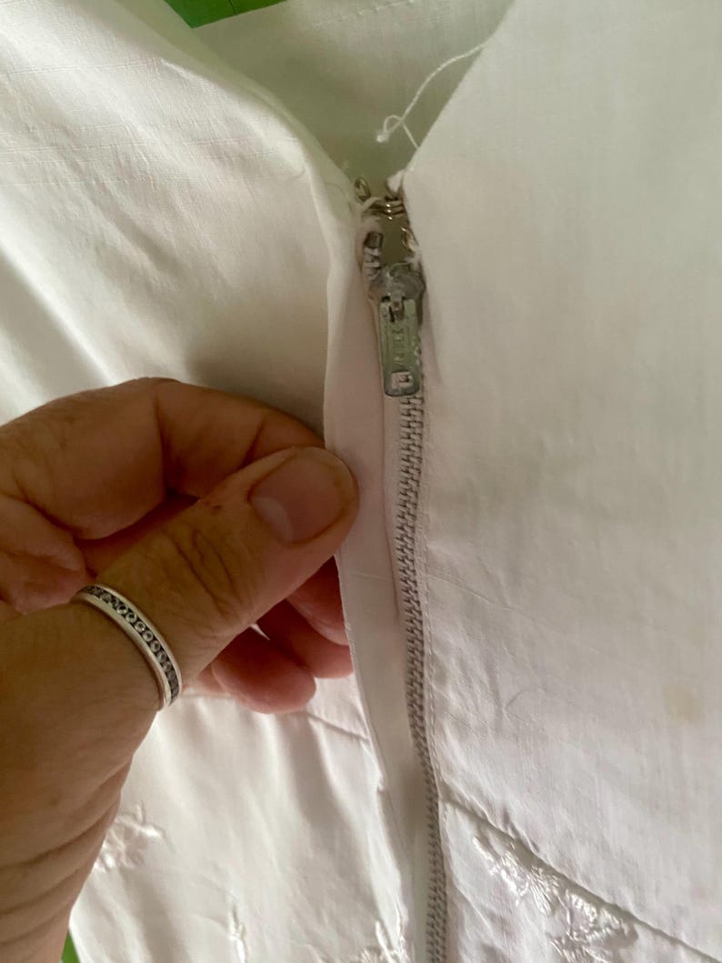 May include: A white garment featuring a vertical metal zip closure with a silver-toned pull tab. The fabric is a solid white material with subtle embroidered details visible near the bottom edge of the closure area.