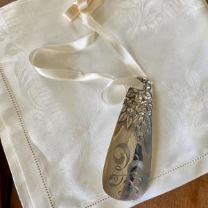 May include: A silver-toned metal shoehorn featuring an embossed floral pattern at the top and an engraved swirling monogram design in the center. A cream-colored satin ribbon is threaded through a hole at the top for hanging purposes.