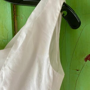 May include: A sleeveless white garment featuring a wide shoulder strap and a clean, smooth fabric texture. The piece is displayed on a black plastic hanger, showcasing the armhole construction and the crisp, solid white colour of the material.