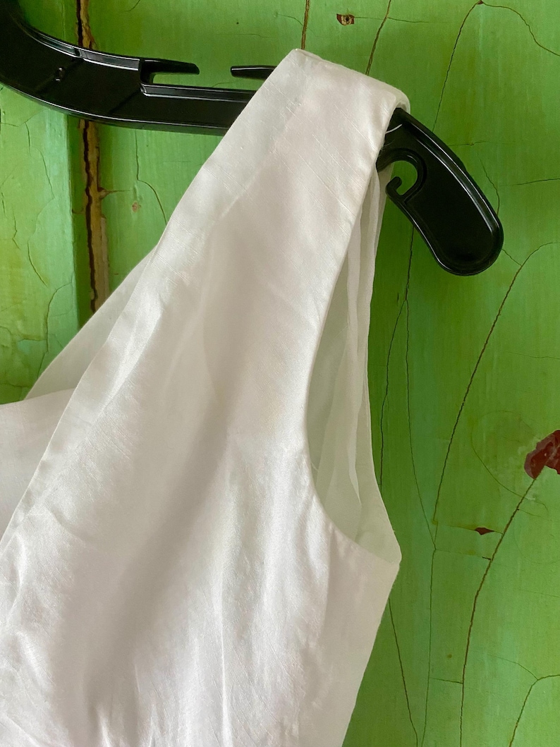 May include: A sleeveless white garment featuring a wide shoulder strap and a clean, smooth fabric texture. The piece is displayed on a black plastic hanger, showcasing the armhole construction and the crisp, solid white colour of the material.