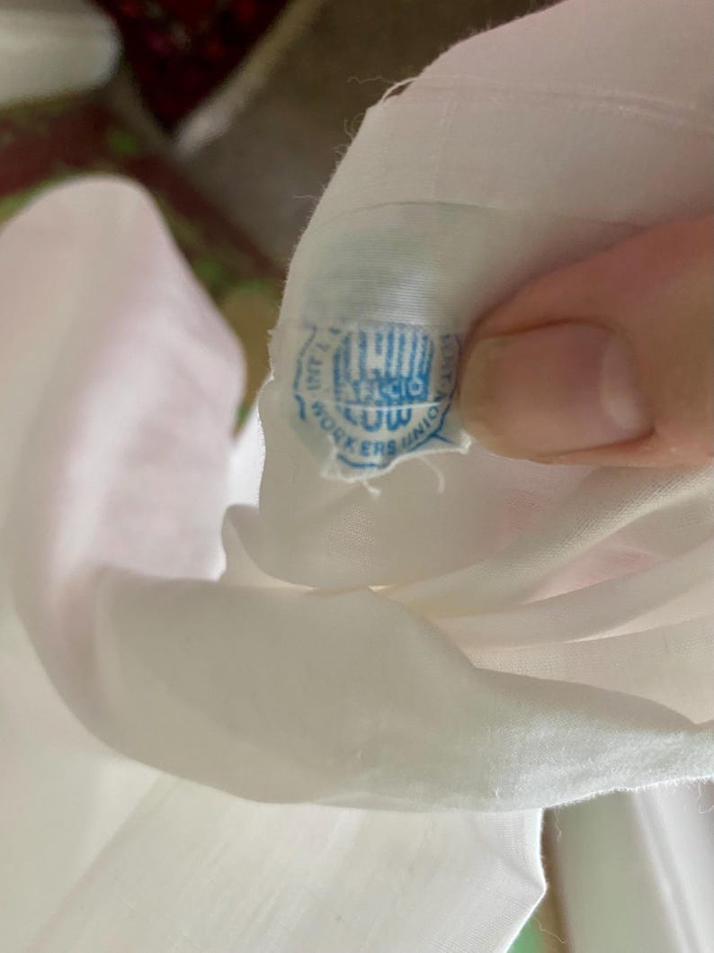 May include: A white garment featuring a small, circular blue and white fabric tag sewn into the interior. The tag displays the text ILGWU, AFL-CIO, and WORKERS UNION in blue ink, indicating the manufacturing origin of the clothing item.